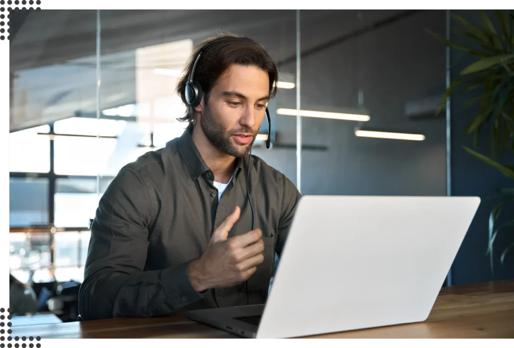 Eine Person sitzt in einem modernen Büro mit Pflanzen und großen Fenstern an einem Laptop und macht erklärende Gestiken während er telefoniert