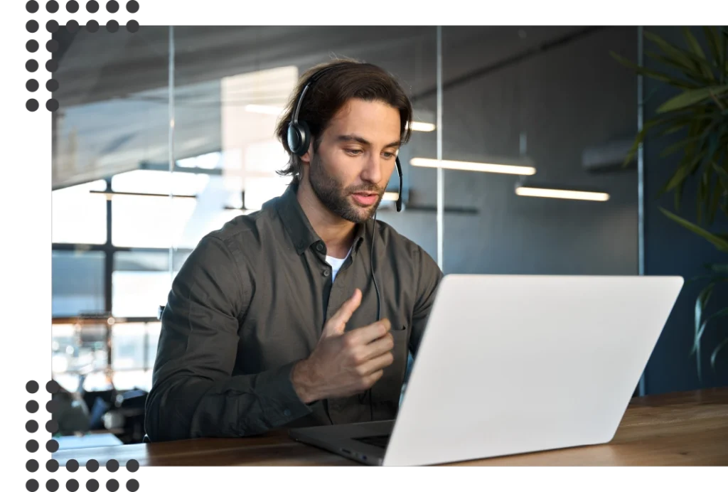 Eine Person sitzt in einem modernen Büro mit Pflanzen und großen Fenstern an einem Laptop und macht erklärende Gestiken während er telefoniert